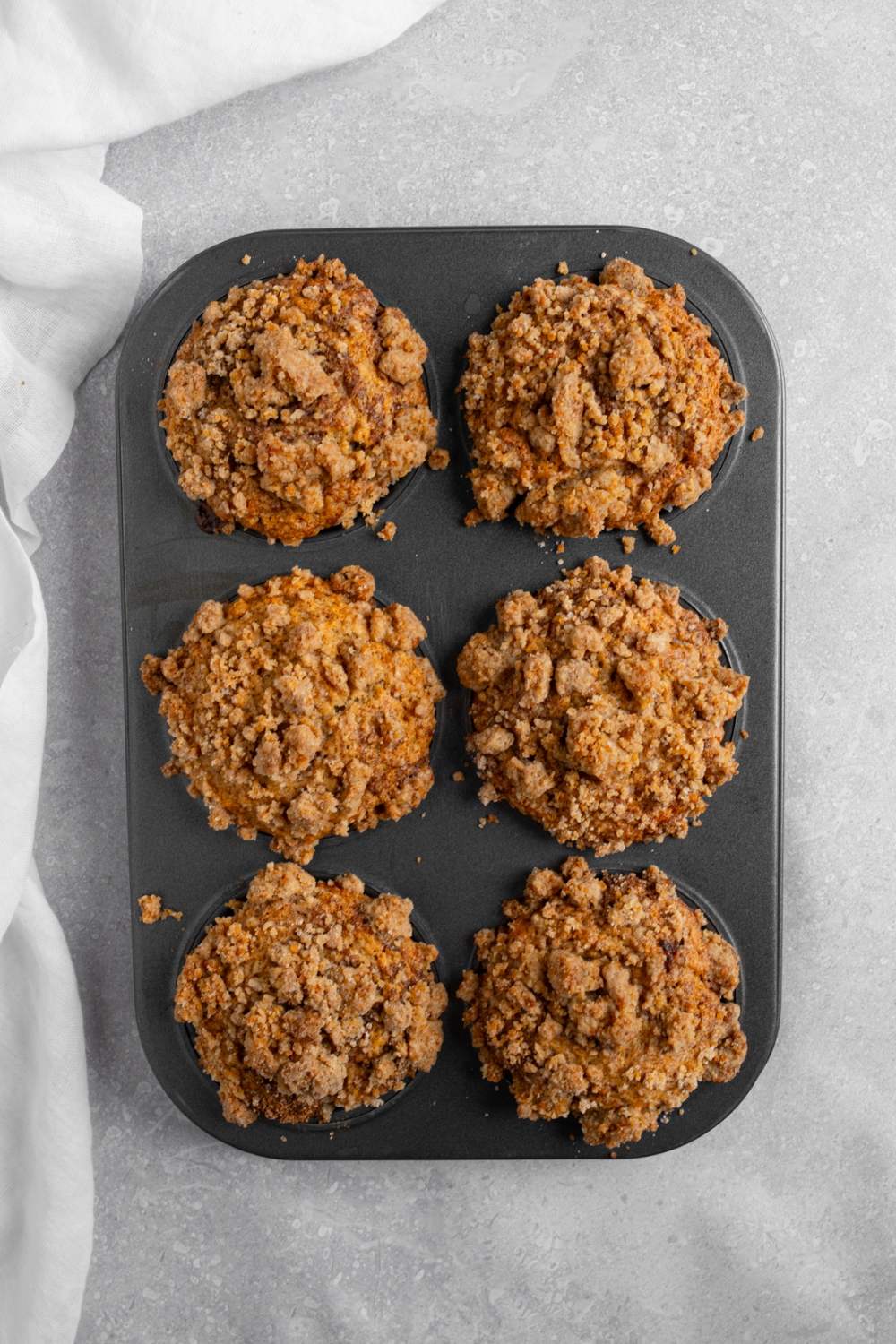 A muffin tin with baked coffee cake muffins. The tray is on a white counter with a white cloth napkin.