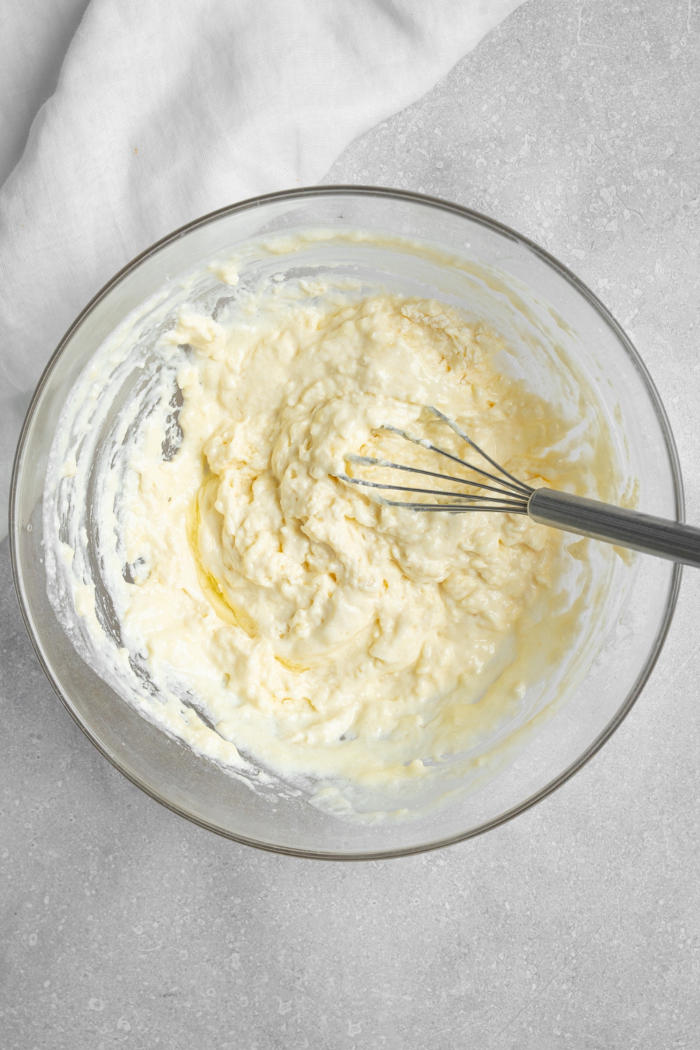 A glass bowl with a whisk stirring pancake batter. The bowl is on a white counter with a white cloth napkin.