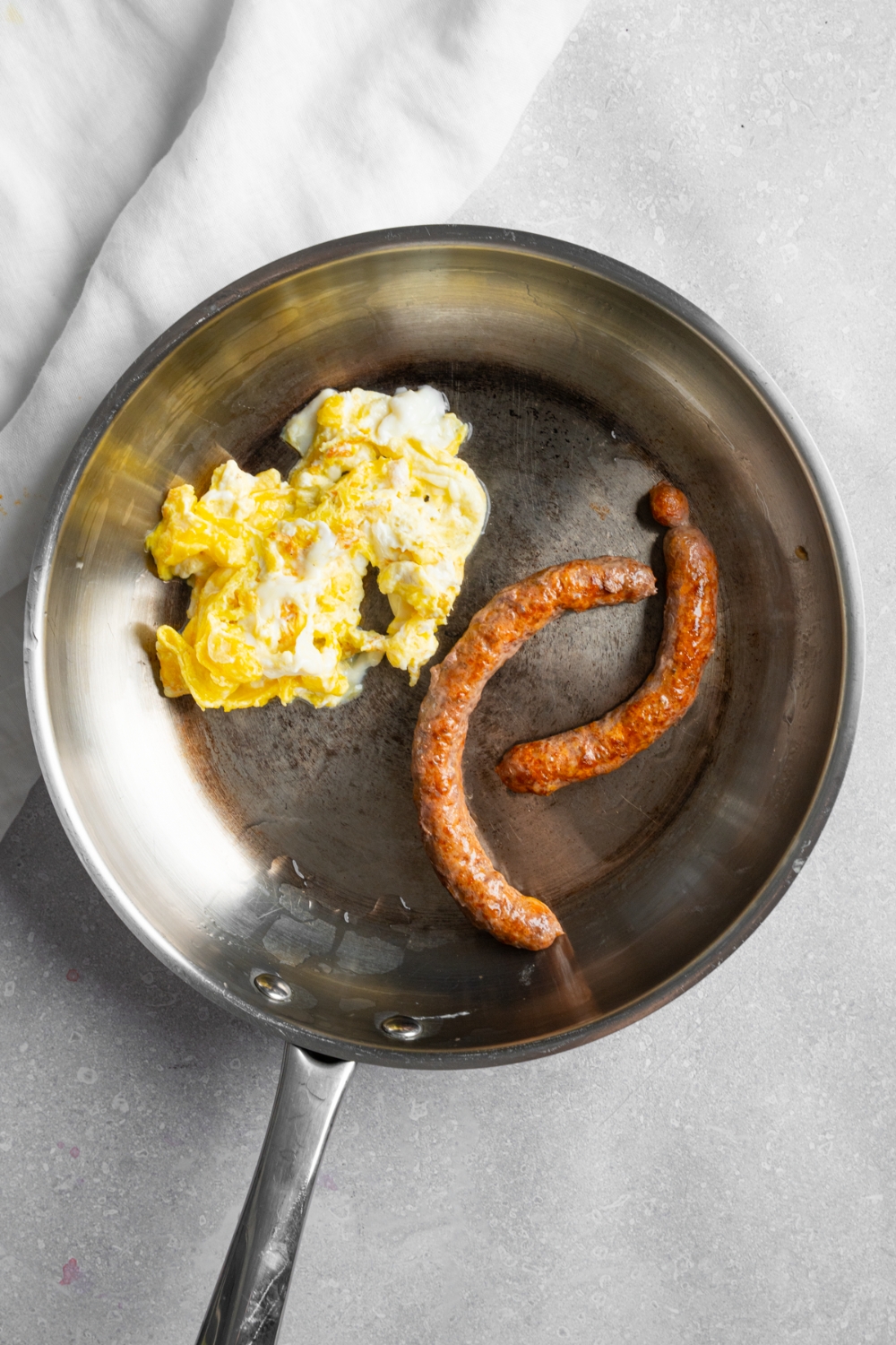 A skillet with scrambled eggs and two cooked sausage links. The skillet is on a white counter with a white cloth napkin.