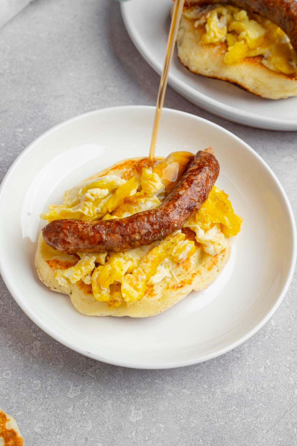 A pancake taco on a white plate with syrup pouring onto the plate. The pancake is layered with scrambled egg and a sausage link drizzled with syrup. The plate is on a white counter with an additional pancake.