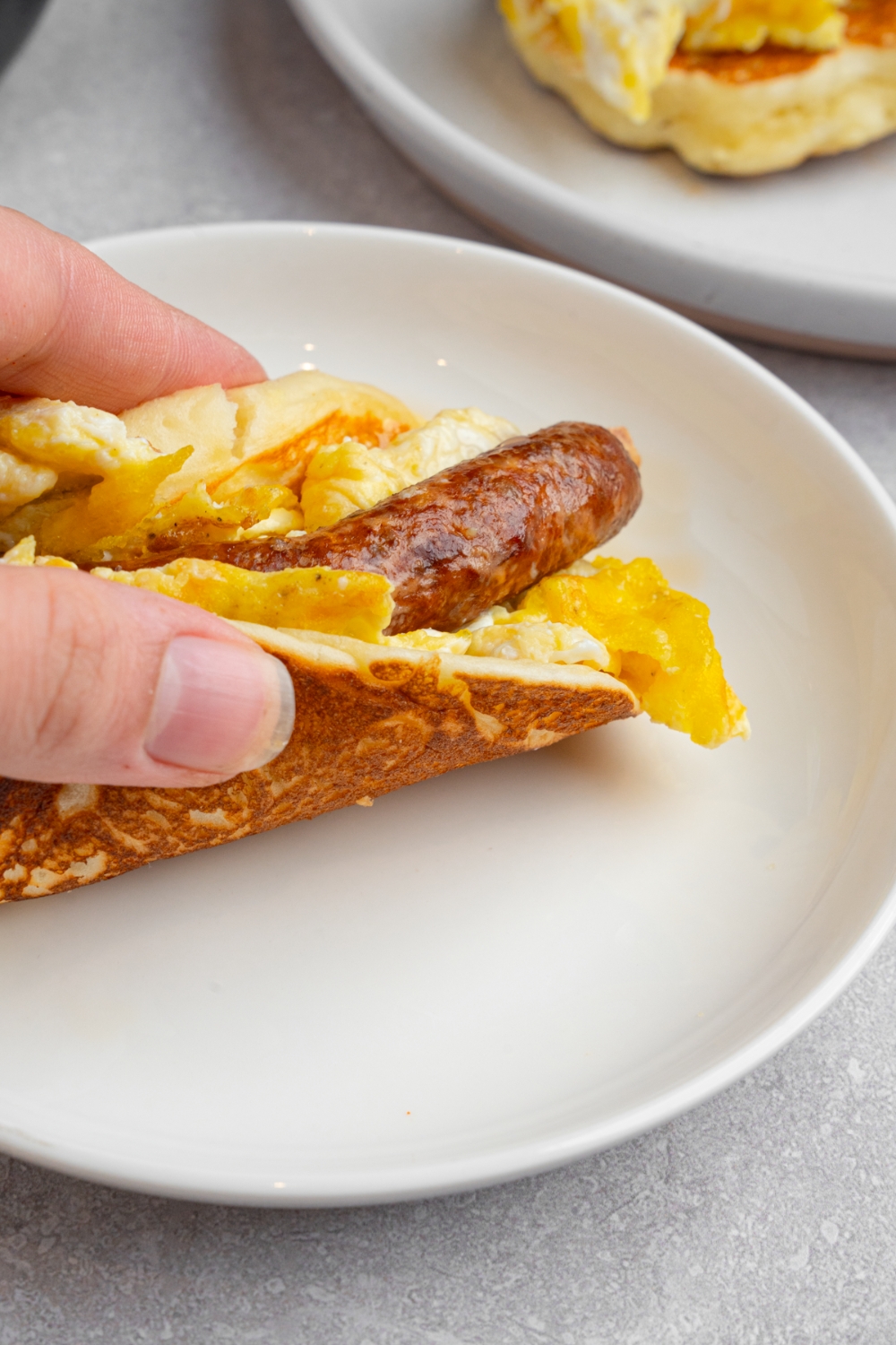 A hand taking a pancake taco from a white plate. The plate is on a white counter with an additional plate with pancakes.