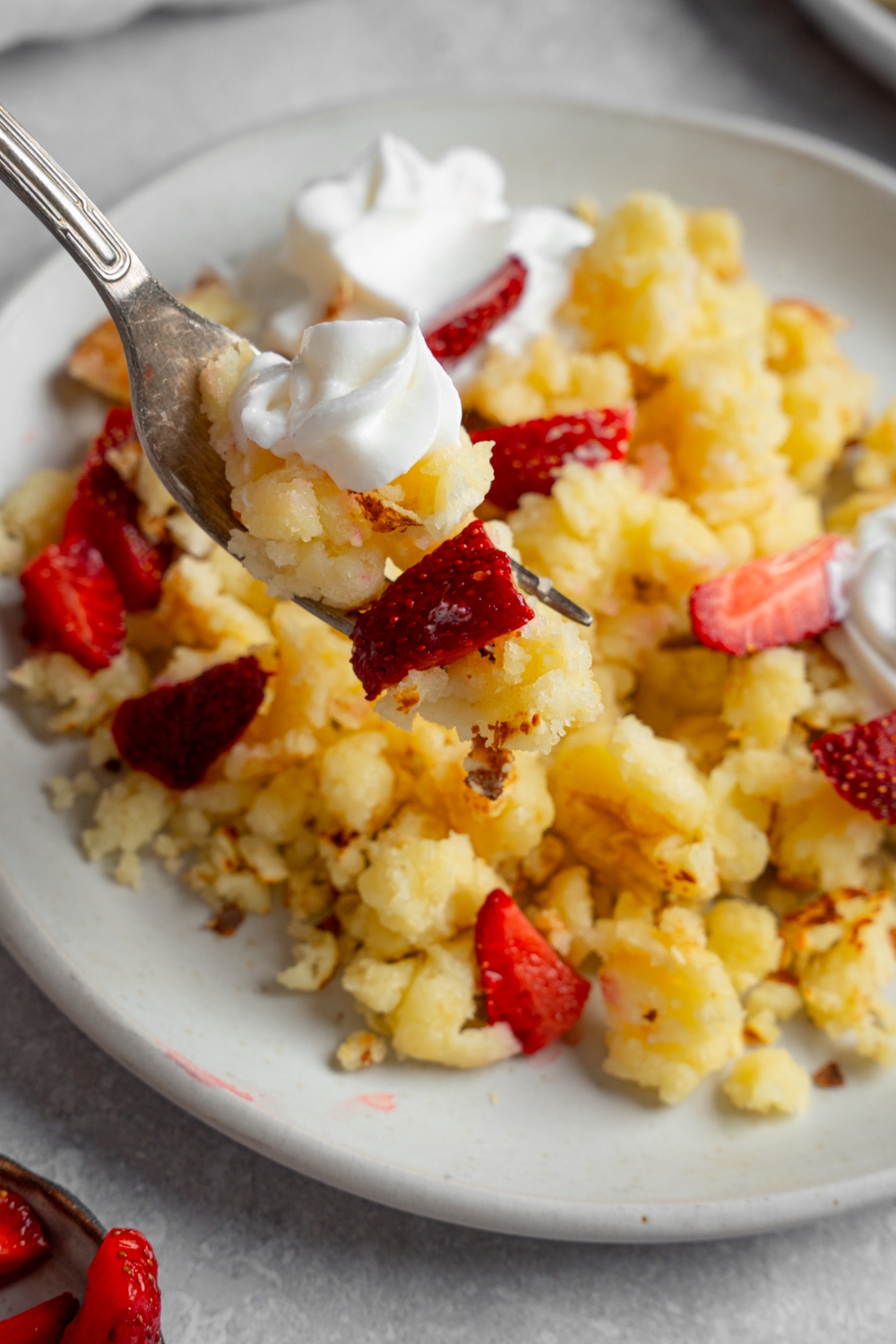 A fork with a bite of scrambled pancakes. There is a plate of scrambled pancakes with strawberries and whipped cream blurred in the background.