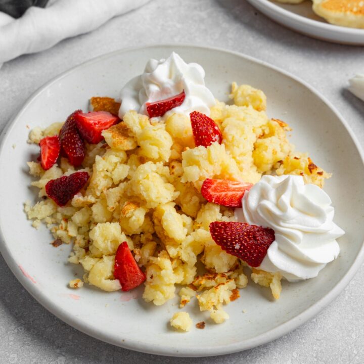 A white plate with scrambled pancakes served with strawberries and whipped cream. The plate is on a white counter with a plate of pancakes and white cloth napkin.