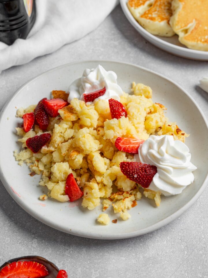 A white plate with scrambled pancakes served with strawberries and whipped cream. The plate is on a white counter with a plate of pancakes and white cloth napkin.