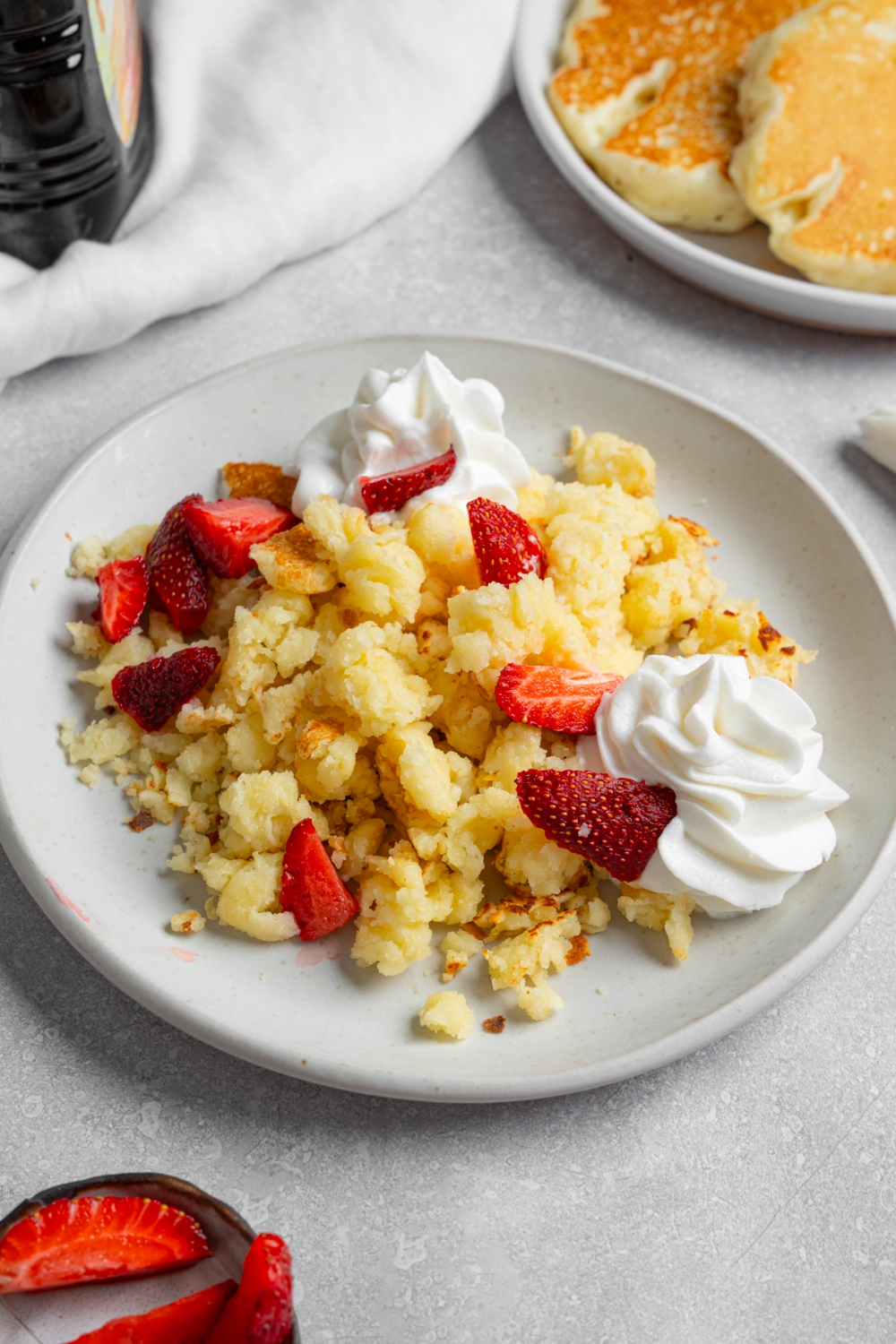 A white plate with scrambled pancakes served with strawberries and whipped cream. The plate is on a white counter with a plate of pancakes and white cloth napkin.