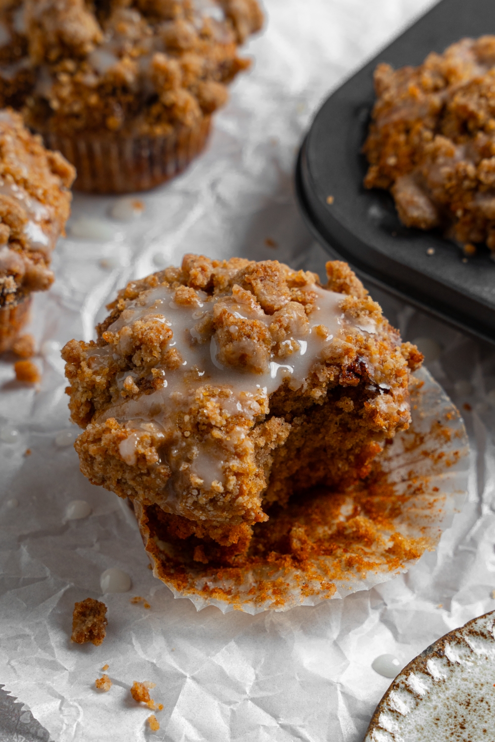 Coffee cake muffins on a sheet of parchment paper with a tray of baked muffins. The muffins are drizzled with a vanilla glaze. There is a bite taken out of one of the muffins.