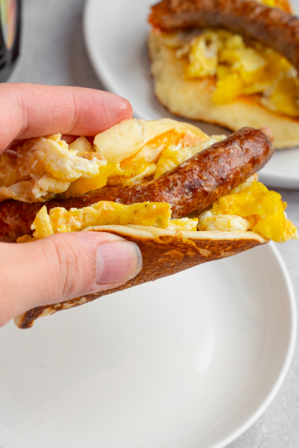 A hand holding a pancake taco. There is a white plate and a plate with an additional pancake taco blurred in the background.
