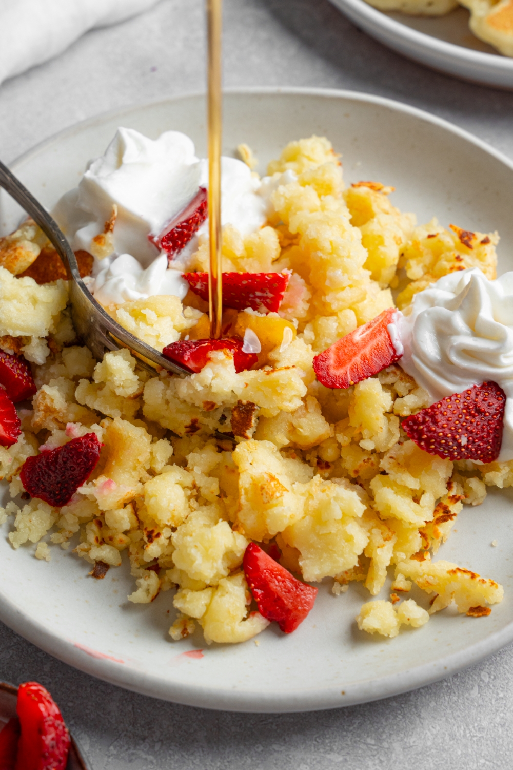 A white plate with scrambled pancakes served with strawberries and whipped cream with syrup being poured on top. A fork is taking a bite of scrambled pancakes.