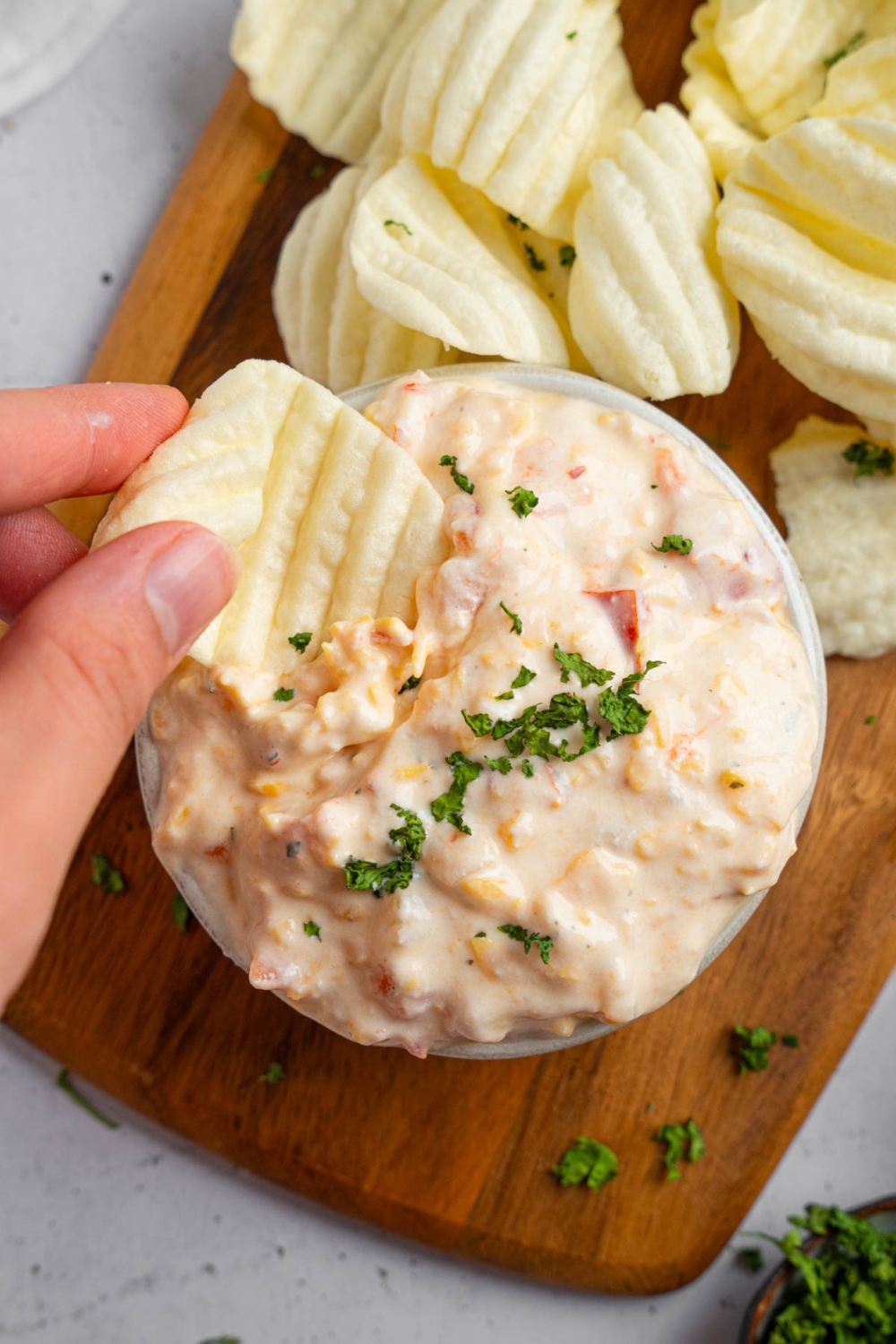 A hand dipping a chip into a bowl of boat dip garnished with chopped parsley. The bowl is on a wooden board with chips.