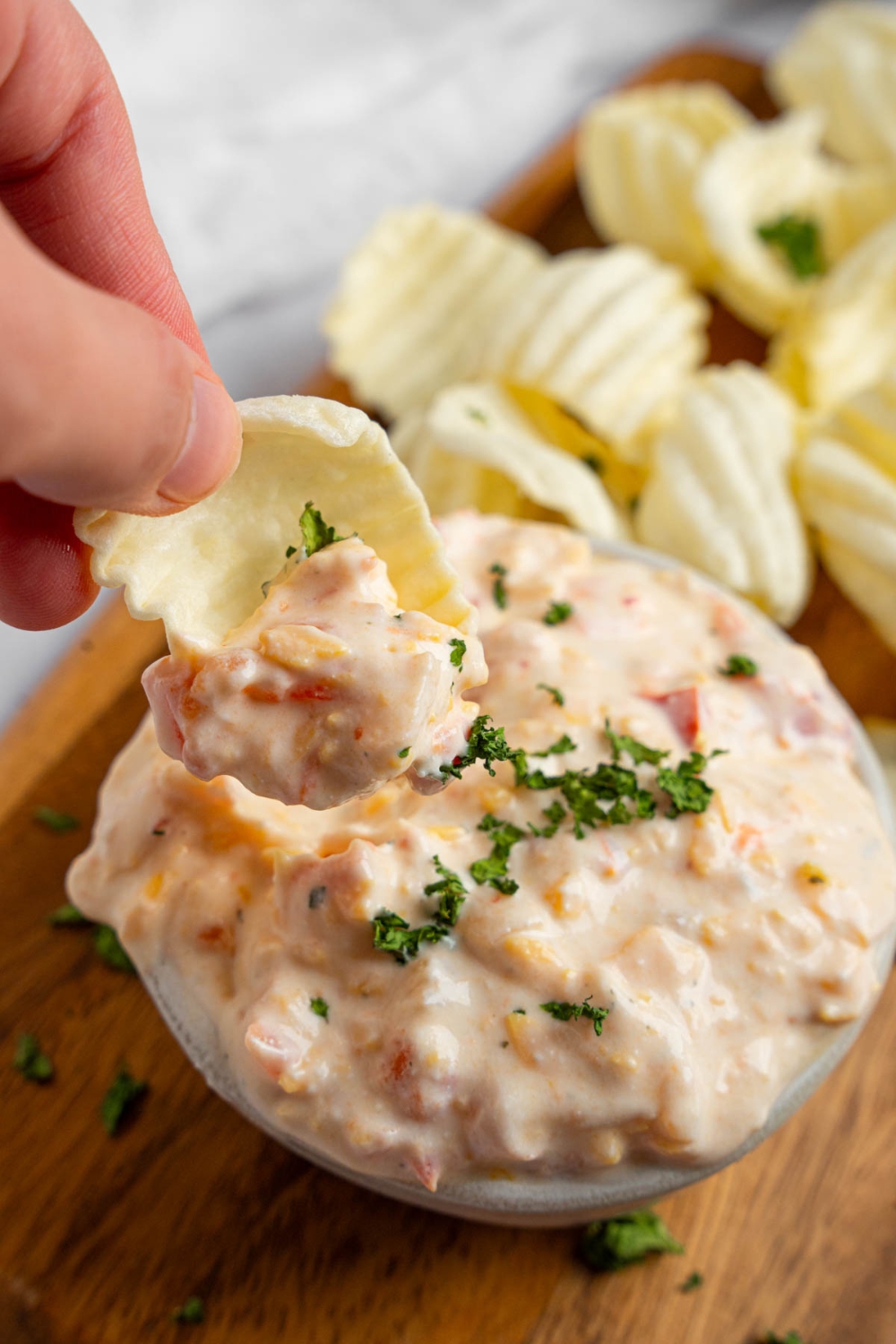 A hand dipping a chip into a bowl of boat dip garnished with chopped parsley. The bowl is on a wooden board with chips.