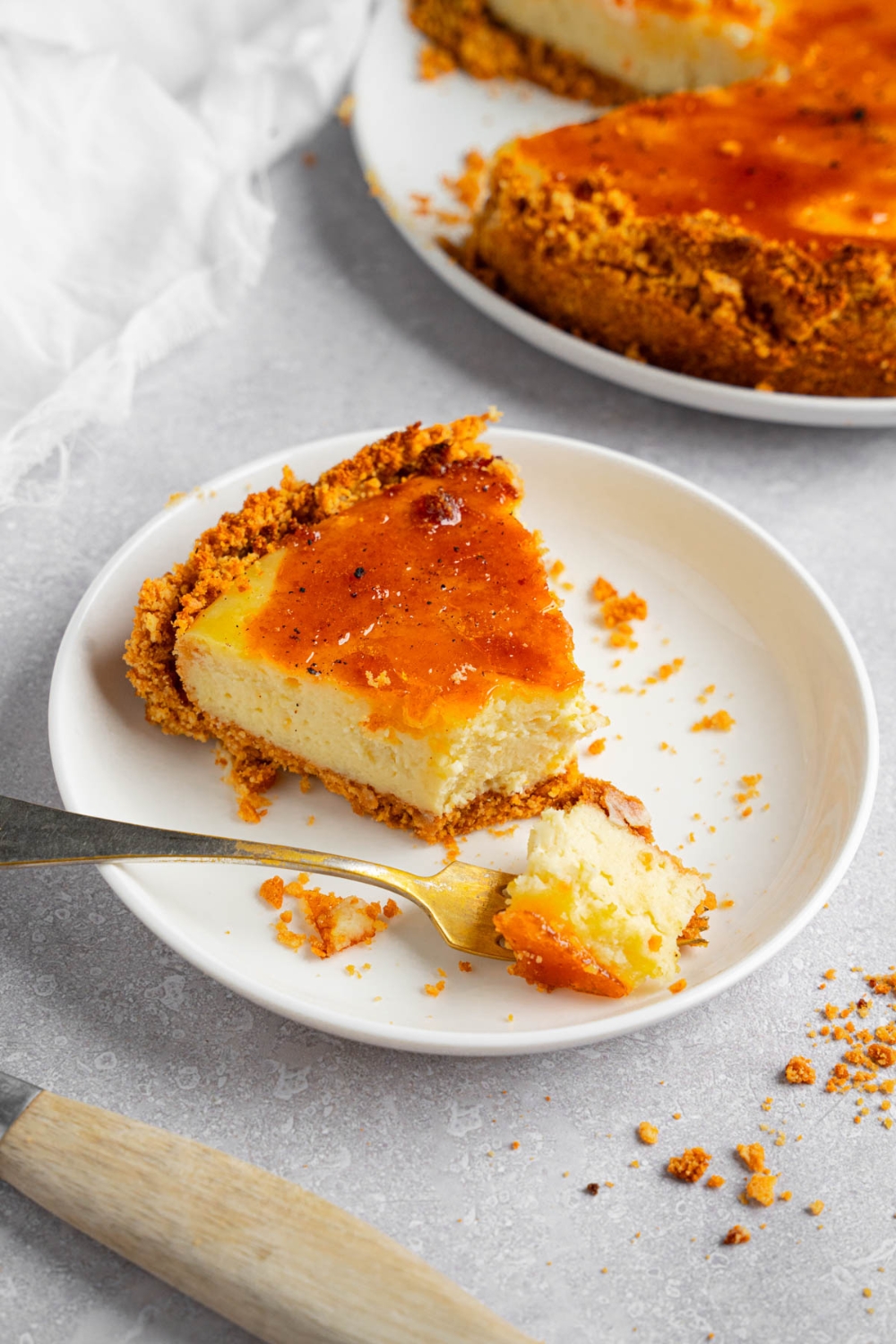 A white plate with a slice of creme brulee cheesecake with a fork with a bite of cheesecake. The plate is on a white counter with the remaining cheesecake and a white cloth napkin.