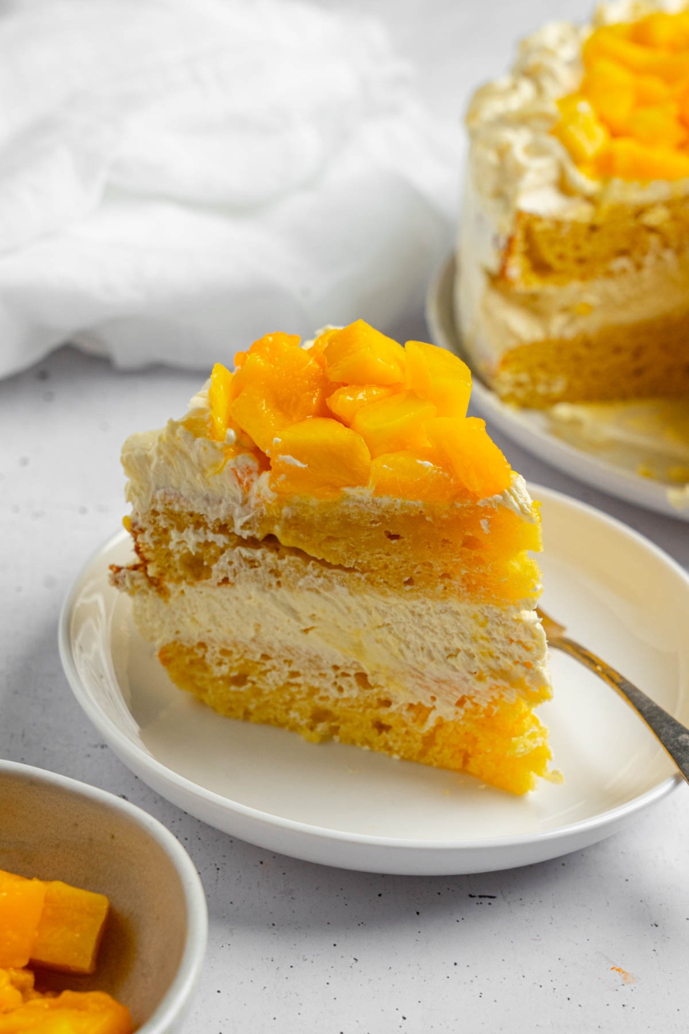 A slice of mango cake on a white plate with a fork. The plate is on a white counter with a whole mango cake and white cloth napkin.