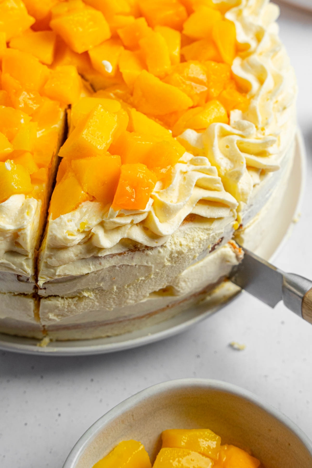A knife cutting a slice of mango cake from a whole cake. The cake is on a white counter with a bowl of cubed mango.