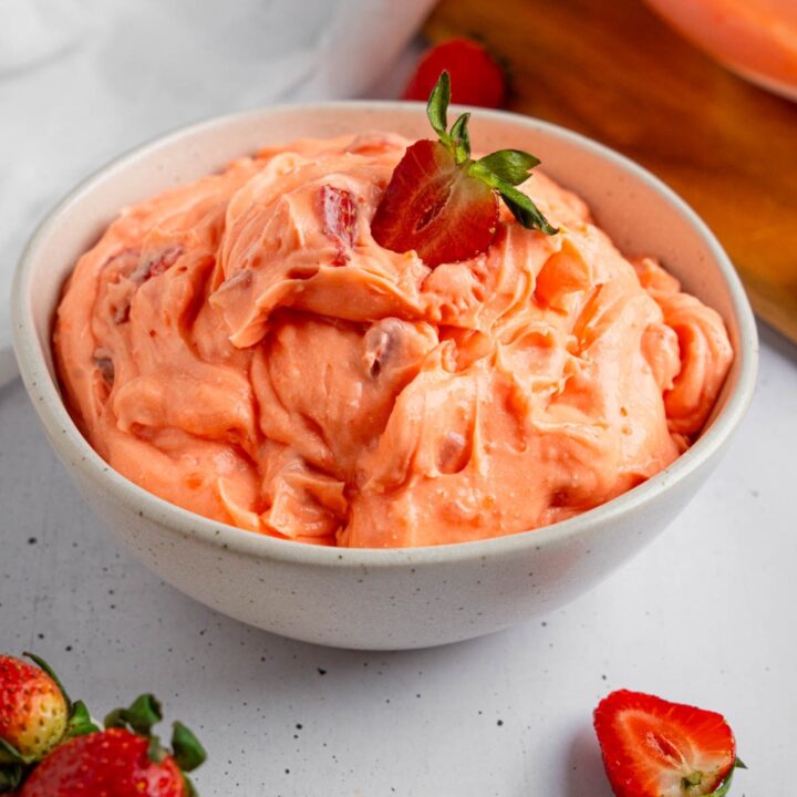 A white bowl of strawberry fluff salad with a sliced strawberry on top. The bowl is on a white counter with strawberries.