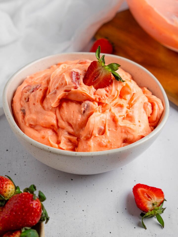 A white bowl of strawberry fluff salad with a sliced strawberry on top. The bowl is on a white counter with strawberries.