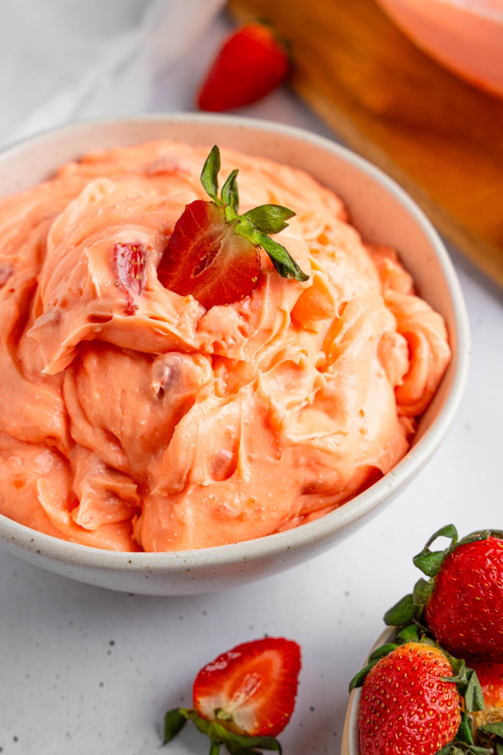 A white bowl of strawberry fluff salad with a sliced strawberry on top. The bowl is on a white counter with strawberries.