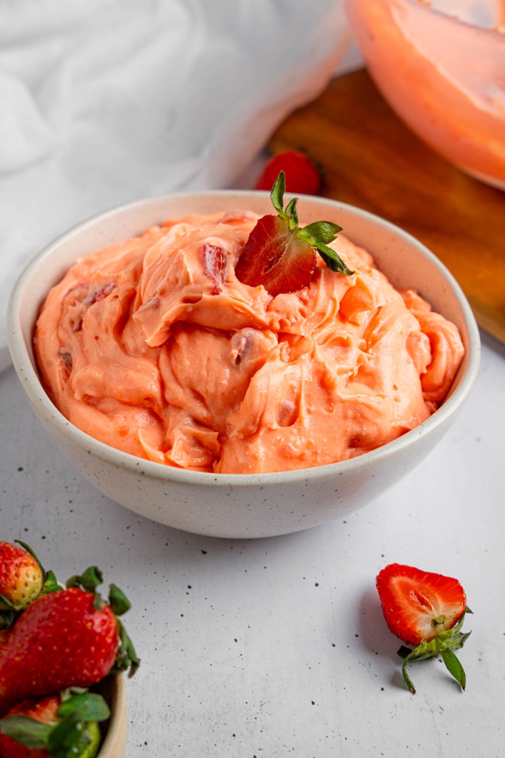 A white bowl of strawberry fluff salad with a sliced strawberry on top. The bowl is on a white counter with strawberries.