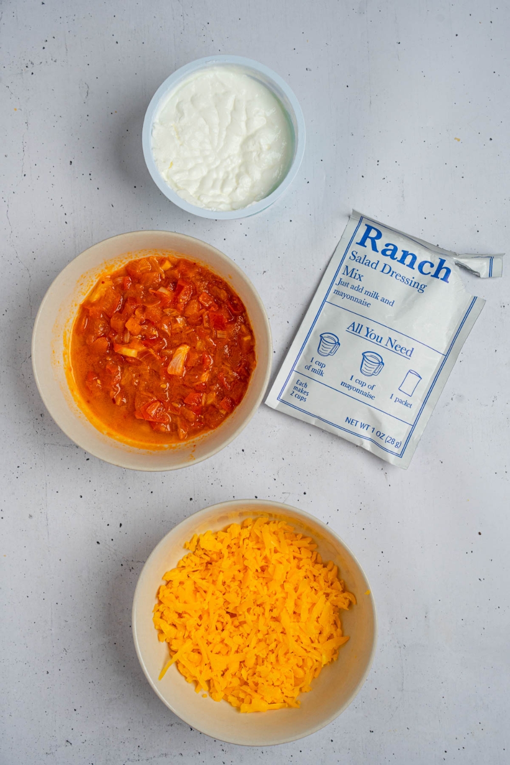 An overhead shot of three bowls containing ingredients to make boat dip including shredded cheddar cheese, sour cream, Rotel, and a packet of spicy Ranch seasoning.
