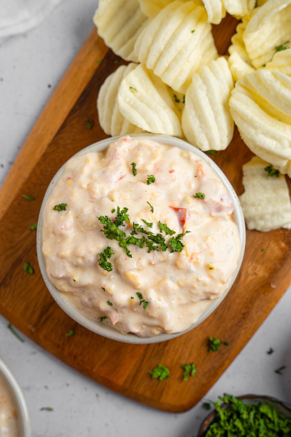 A bowl of boat dip garnished with chopped parsley. The bowl is on a wooden board with chips. The board is on a white counter with a ramekin of garnishes.