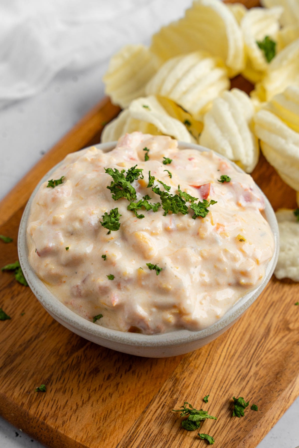 A bowl of boat dip garnished with chopped parsley. The bowl is on a wooden board with chips. The board is on a white counter.