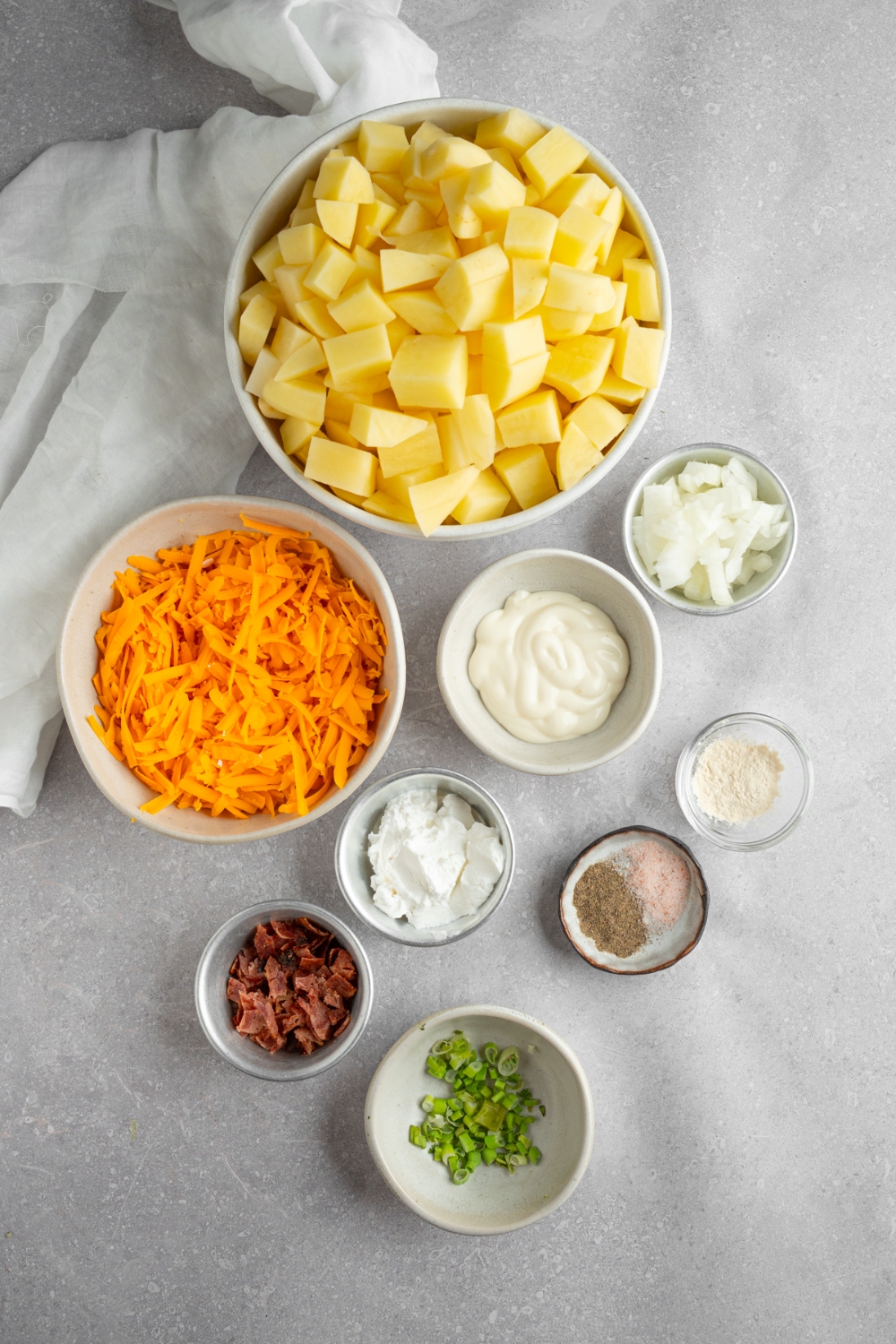 An overhead shot of several bowls in various sizes containing ingredients to make Mississippi mud potatoes including shredded cheddar cheese, cubed potatoes, sour cream, mayo, onion, bacon, a and seasonings.