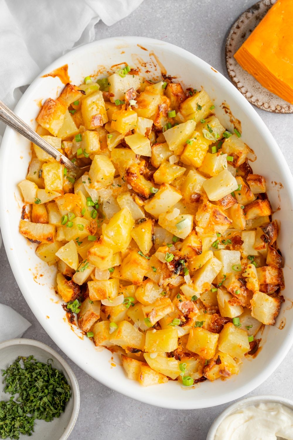 A baking dish of cheesy Mississippi mud potatoes garnished with sliced green onion. The dish is on a white counter with a spoon in the potatoes. The dish is next to a ramekin of garnishes and a brick of cheddar cheese.