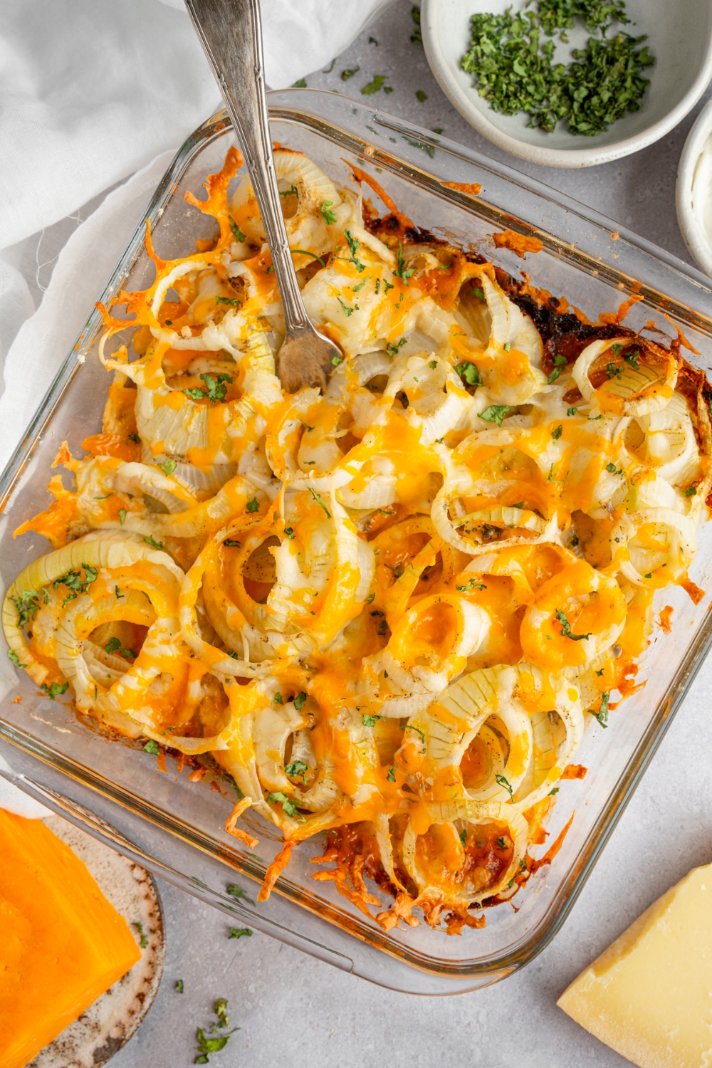 A square glass baking dish with Tennessee onions garnished with fresh parsley with a fork in the onions. The dish is on a white counter with a small bowl of garnishes and brick of cheese.