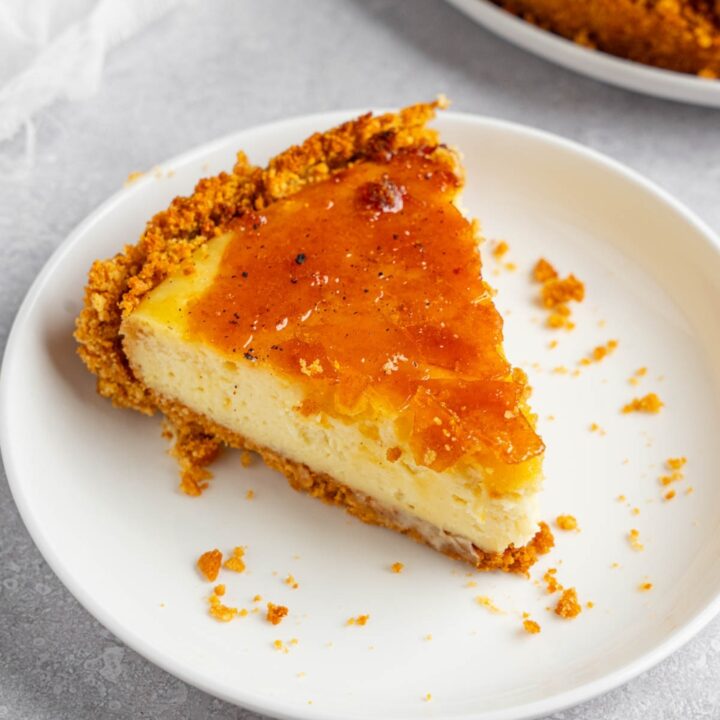 A white plate with a slice of creme brulee cheesecake. The plate is on a white counter with the remaining cheesecake and a white cloth napkin.