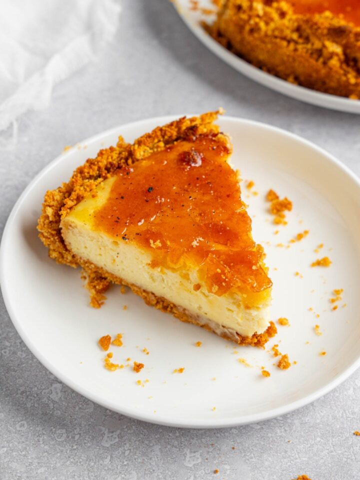 A white plate with a slice of creme brulee cheesecake. The plate is on a white counter with the remaining cheesecake and a white cloth napkin.