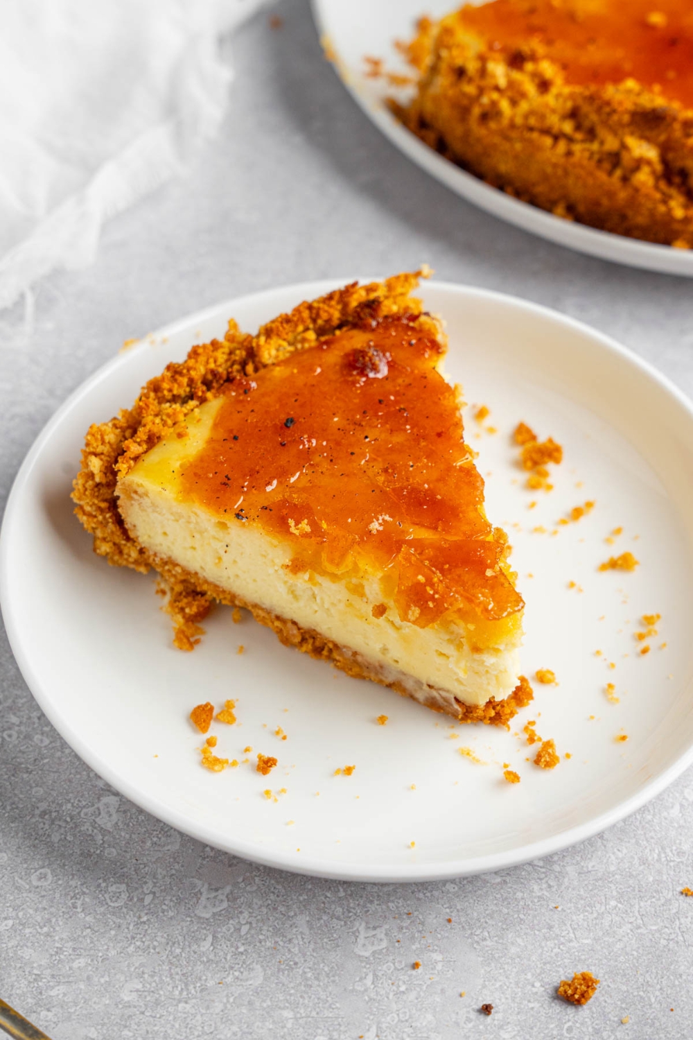 A white plate with a slice of creme brulee cheesecake. The plate is on a white counter with the remaining cheesecake and a white cloth napkin.
