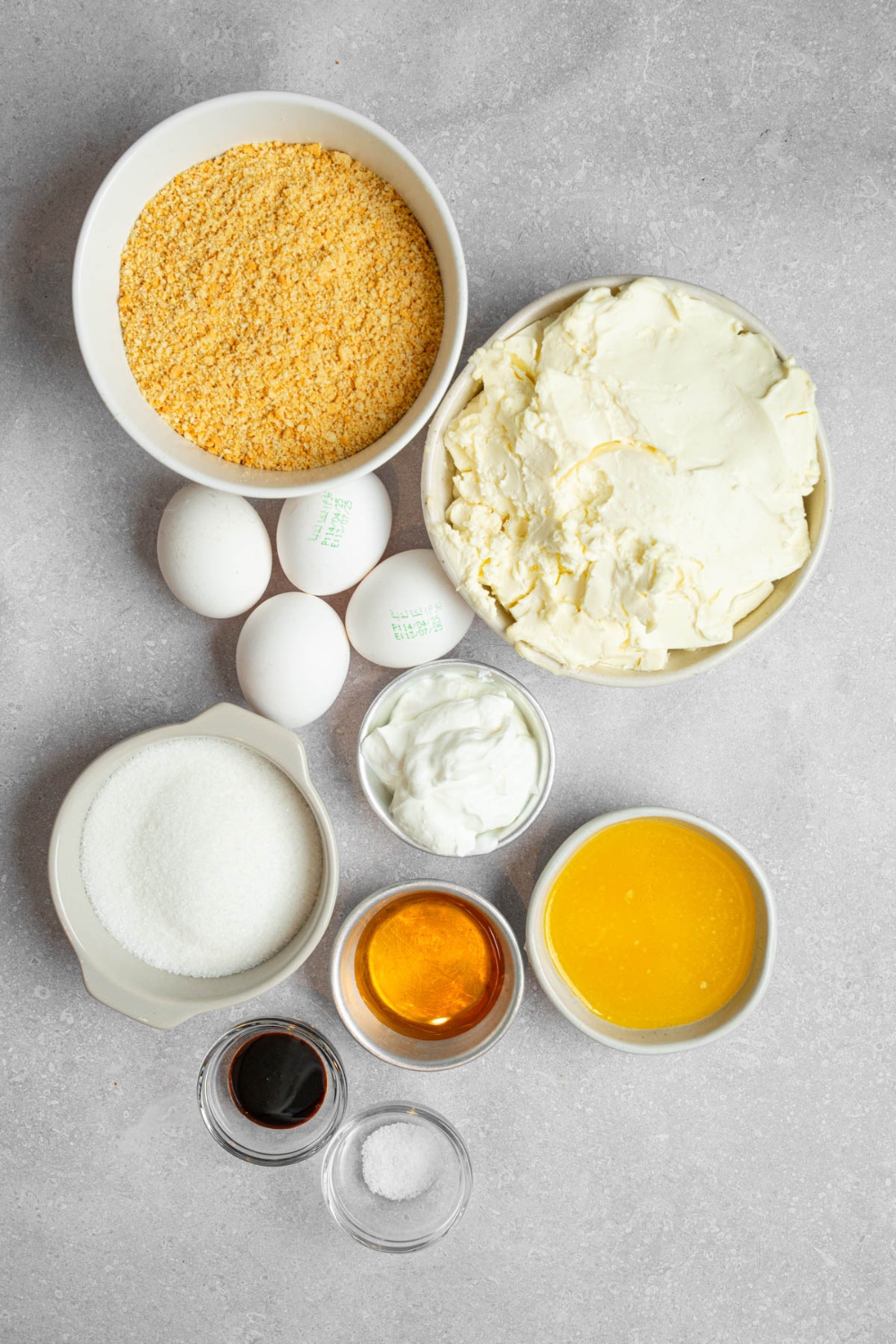 An overhead shot of several bowls in various sizes containing ingredients to make creme brulee cheesecake including cream cheese, graham cracker crumbs, eggs, vanilla, sugar, sour cream, butter, and honey.