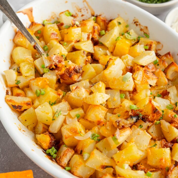 A baking dish of cheesy Mississippi mud potatoes garnished with sliced green onion. The dish is on a white counter with a spoon in the potatoes. The dish is next to a ramekin of garnishes and a brick of cheddar cheese.