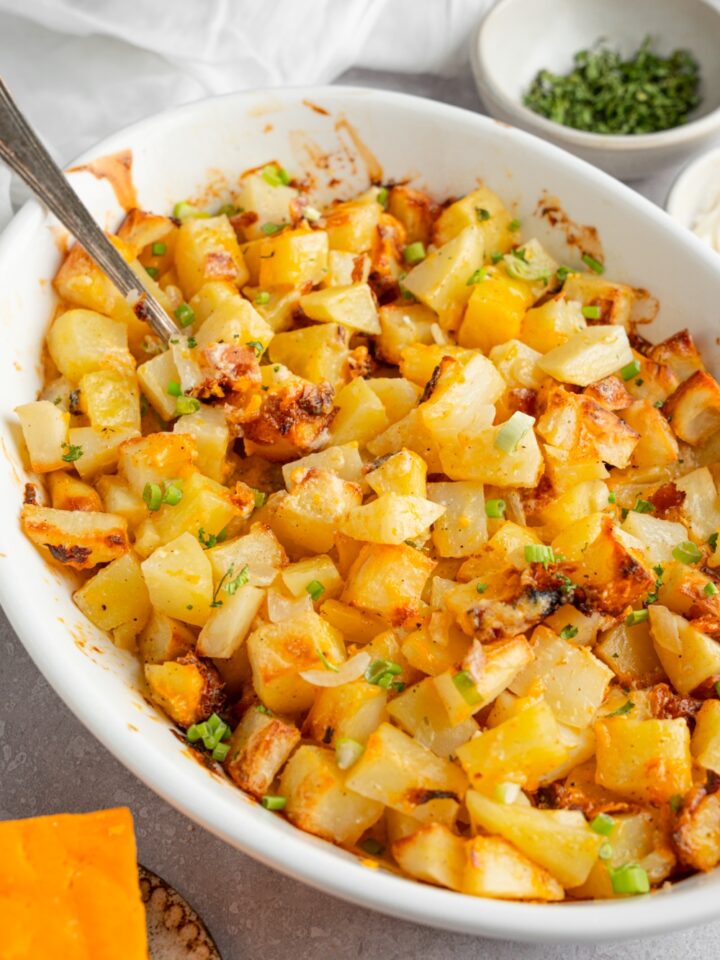 A baking dish of cheesy Mississippi mud potatoes garnished with sliced green onion. The dish is on a white counter with a spoon in the potatoes. The dish is next to a ramekin of garnishes and a brick of cheddar cheese.