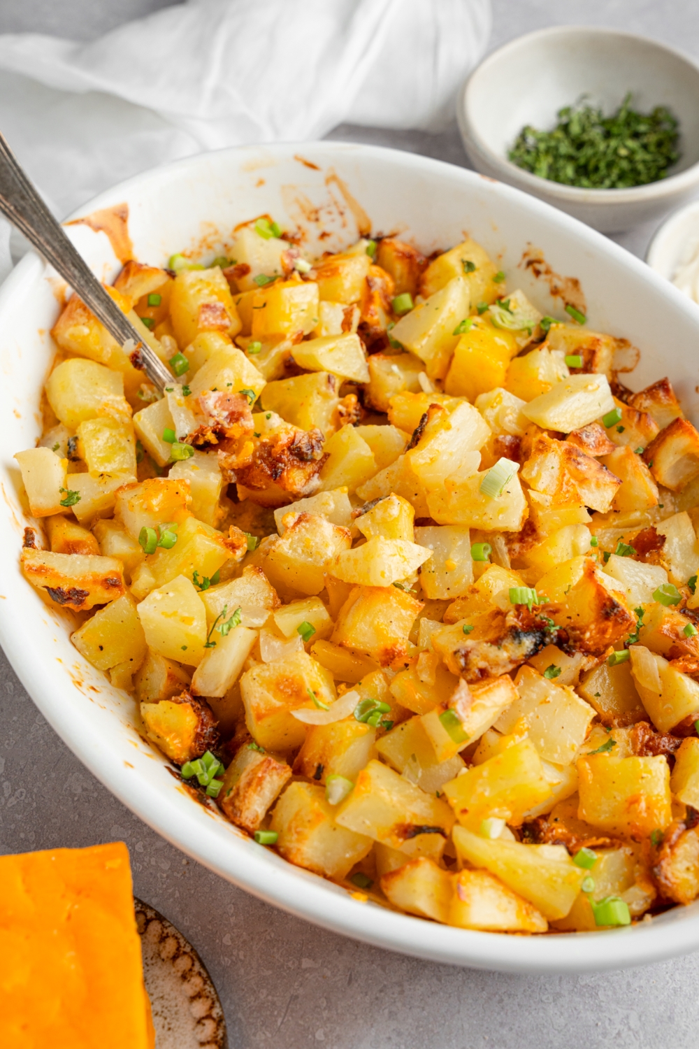 A baking dish of cheesy Mississippi mud potatoes garnished with sliced green onion. The dish is on a white counter with a spoon in the potatoes. The dish is next to a ramekin of garnishes and a brick of cheddar cheese.