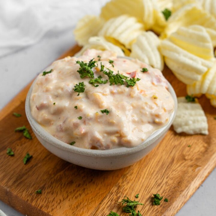 A bowl of boat dip garnished with chopped parsley. The bowl is on a wooden board with chips. The board is on a white counter with a ramekin of garnishes.