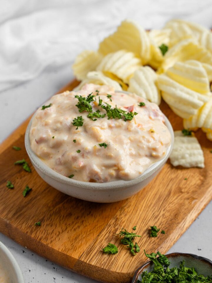 A bowl of boat dip garnished with chopped parsley. The bowl is on a wooden board with chips. The board is on a white counter with a ramekin of garnishes.