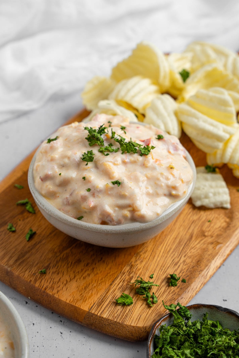 A bowl of boat dip garnished with chopped parsley. The bowl is on a wooden board with chips. The board is on a white counter with a ramekin of garnishes.