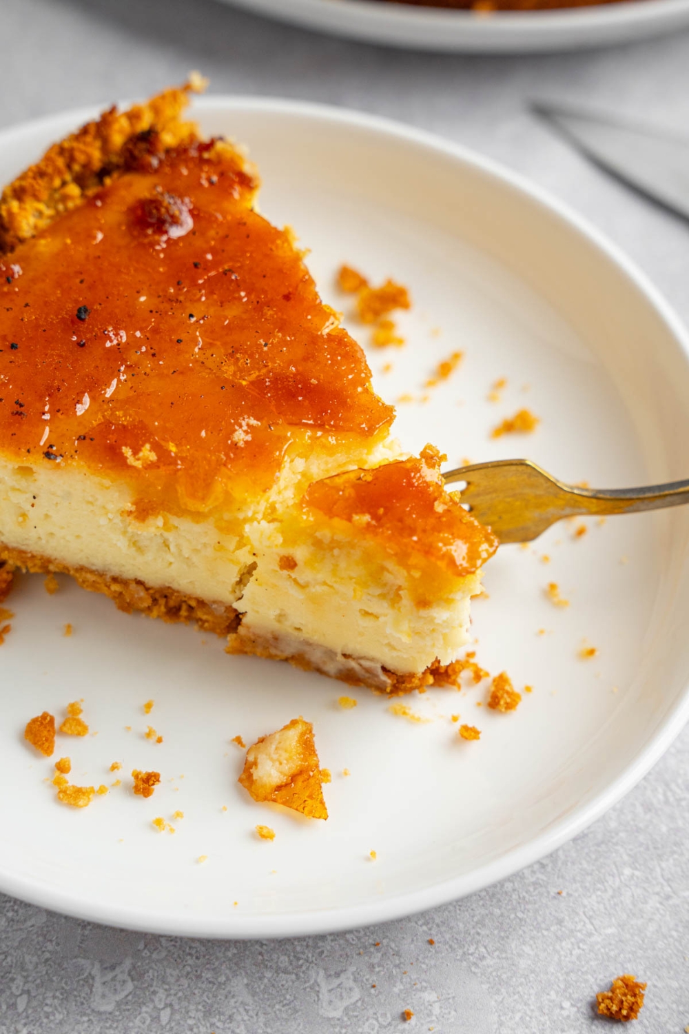 A white plate with a slice of creme brulee cheesecake with a fork with a bite of cheesecake. The plate is on a white counter with a white cloth napkin.