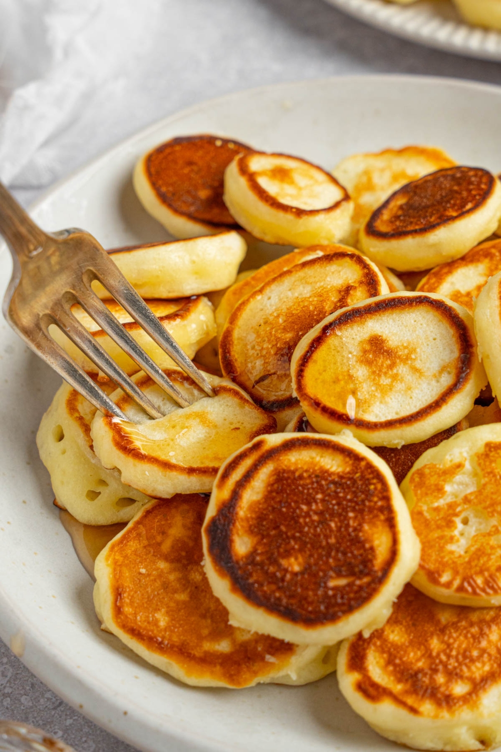 A fork taking a bite of mini pancakes from a white plate of pancakes. The plate is on a white counter.