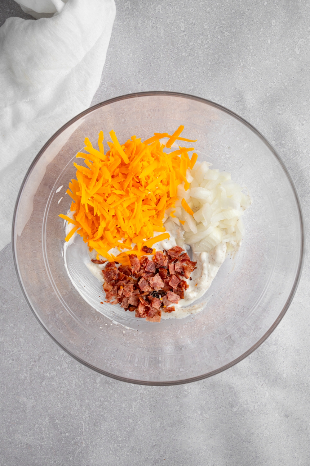A glass bowl with mayo, sour cream, onion, shredded cheddar cheese, and bacon. The bowl is on a white counter with a white cloth napkin.