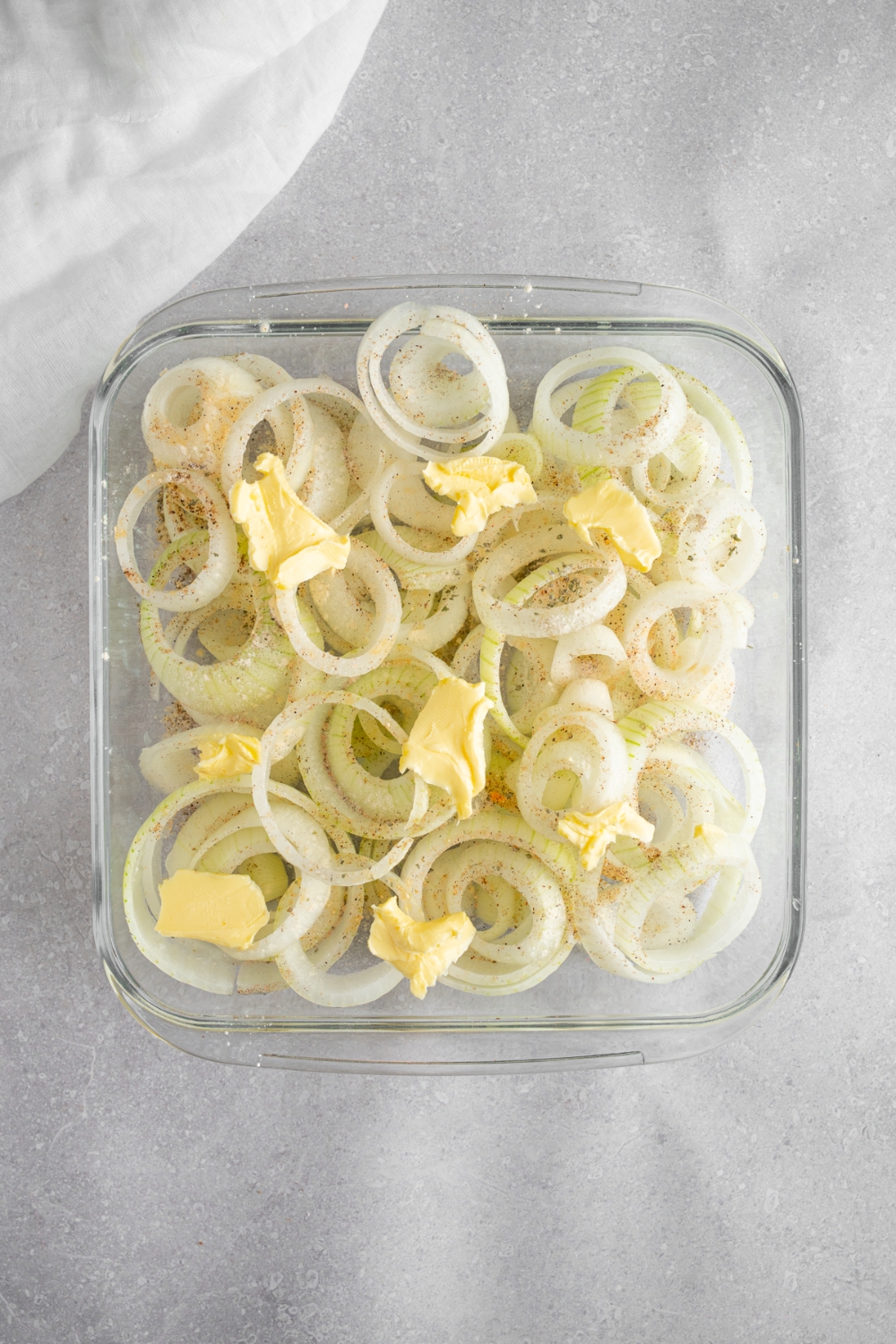 A square glass baking dish with sliced onion rings topped with butter and seasonings. The dish is on a white counter with a white cloth napkin.