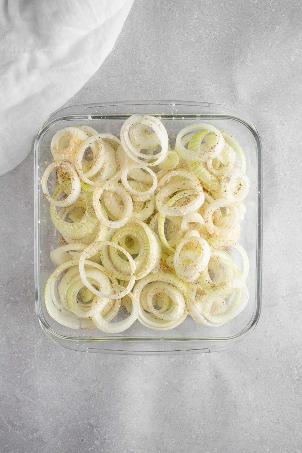 A square glass baking dish with sliced onion rings topped with seasonings. The dish is on a white counter with a white cloth napkin.