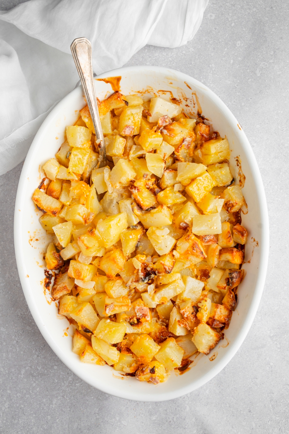 A baking dish with cooked cheesy Mississippi mud potatoes with a spoon in the potatoes. The dish is on a white counter with a white cloth napkin.