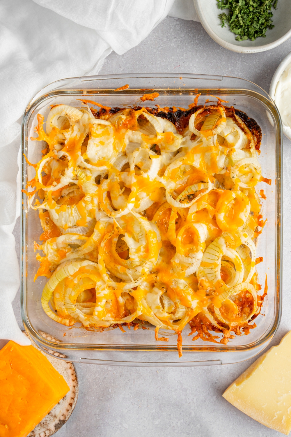 A square glass baking dish with cooked Tennessee onions. The dish is on a white counter with a small ramekin of garnishes and a brick of cheese.