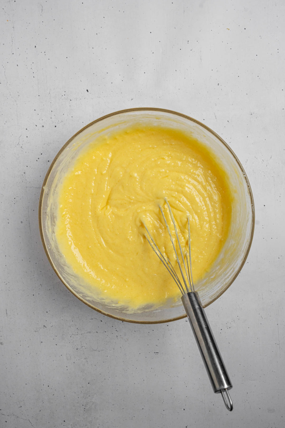 A glass bowl with a whisk mixing mango cake batter. The bowl is on a white counter.