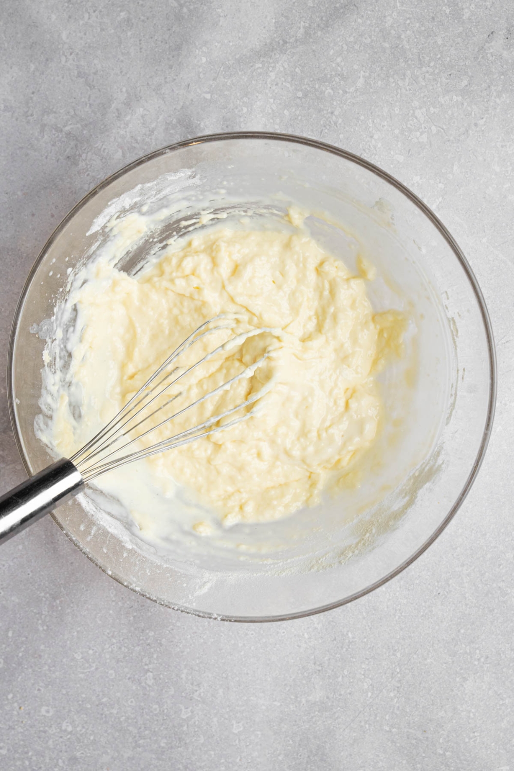 A glass bowl with a whisk stirring mini pancake batter. The bowl is on a white counter.