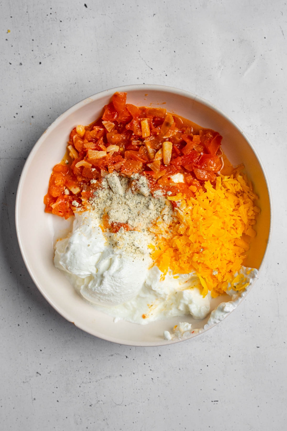 A white bowl with ingredients to make boat dip including shredded cheddar cheese, mild Rotel, sour cream, and spicy ranch seasoning. The bowl is on a white counter.
