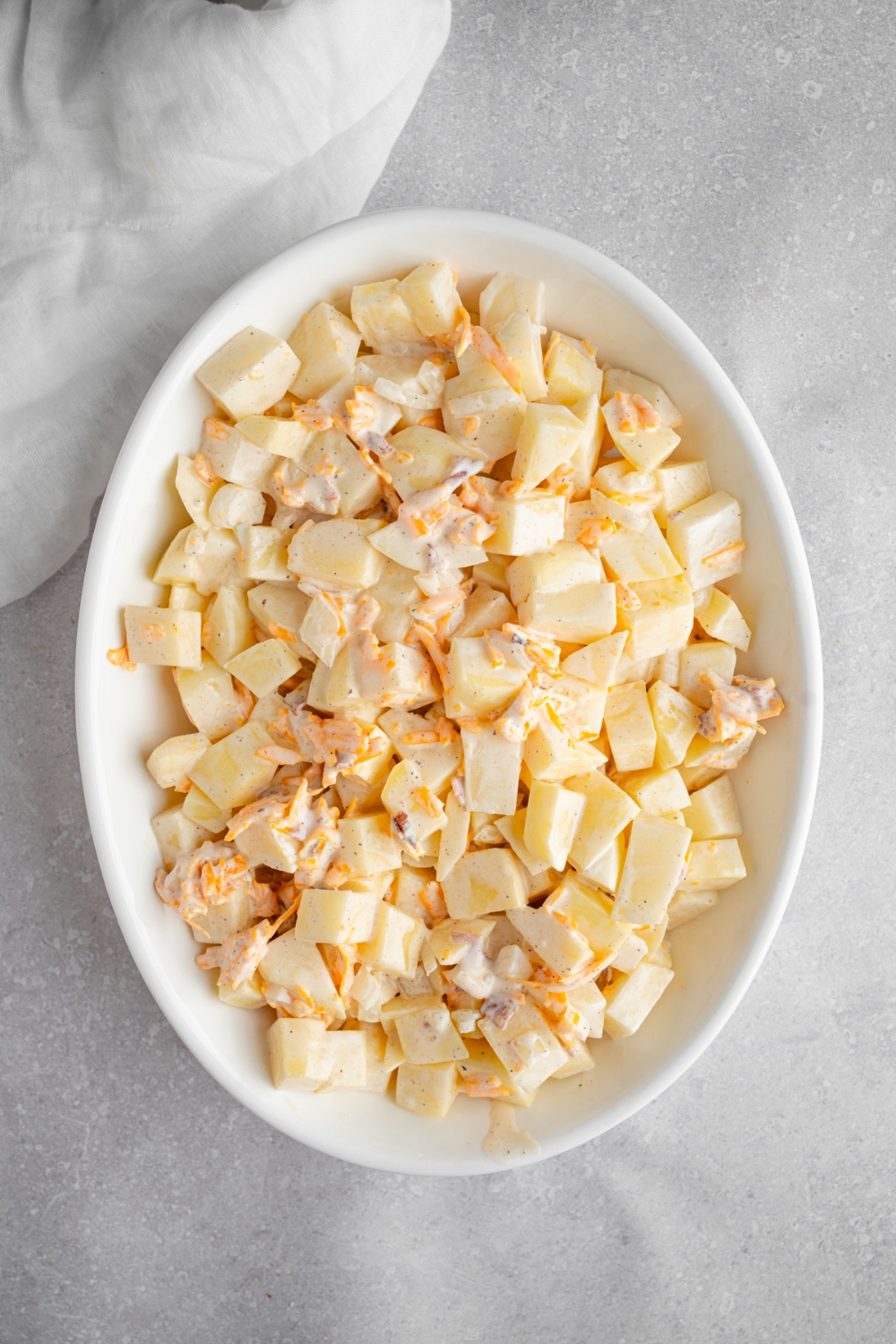 A baking dish with uncooked potatoes tossed in a creamy mayo and cheese mixture. The dish is on a white counter with a white cloth napkin.