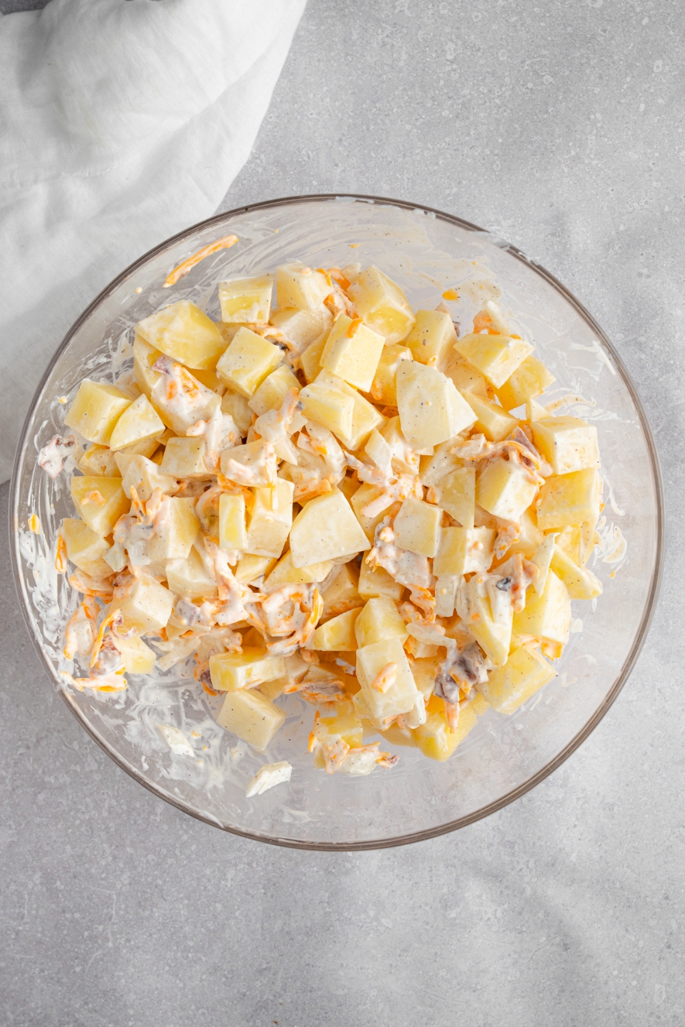 A glass bowl with cubed potatoes tossed in a mayo and cheddar cheese mixture. The bowl is on a white counter with a white cloth napkin.