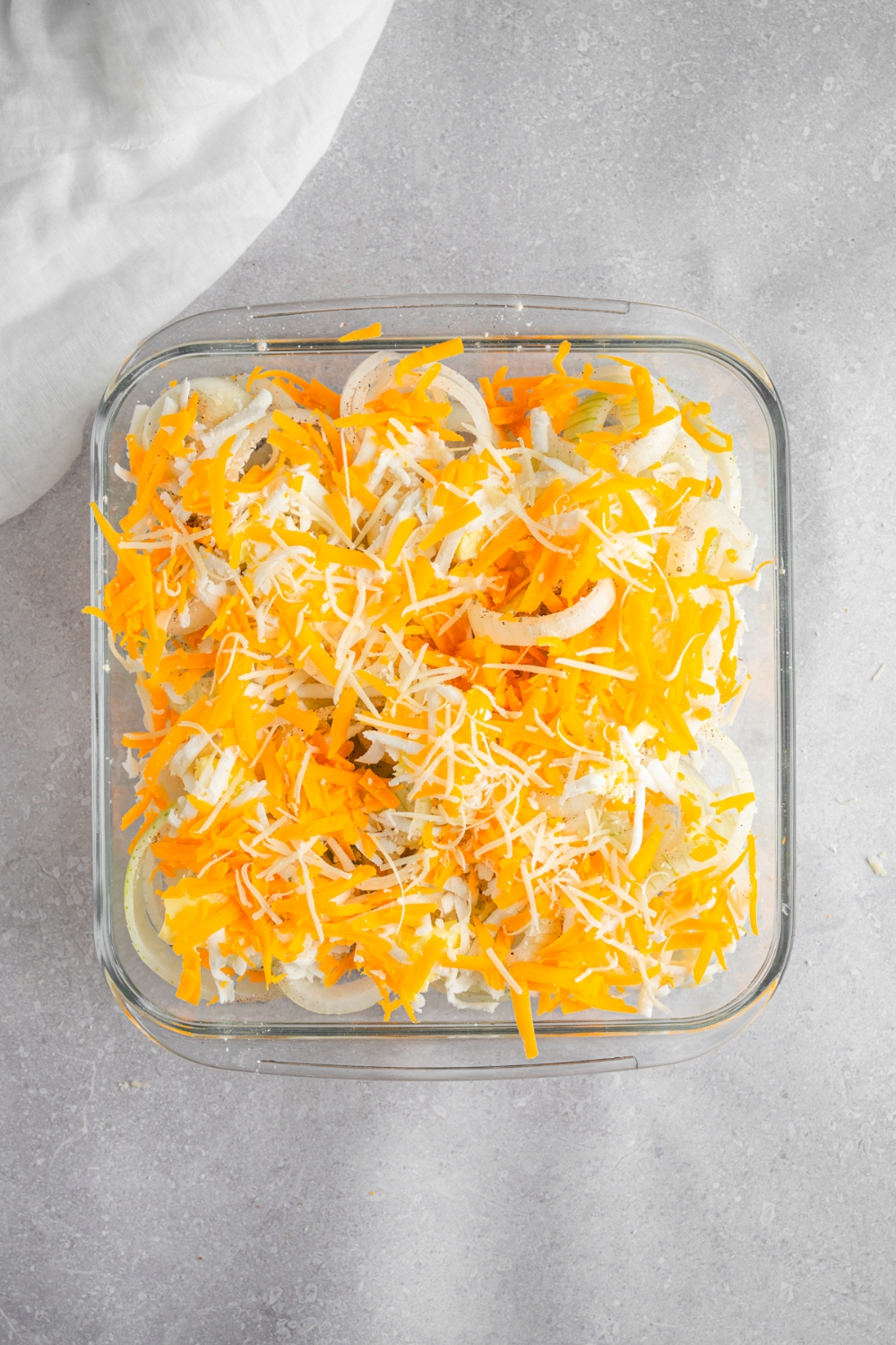 A square glass baking dish with sliced onion rings topped with shredded cheeses. The dish is on a white counter with a white cloth napkin.